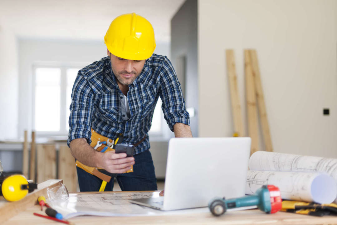 Construction worker looking at his phone and laptop while on the job.